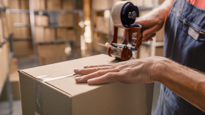 Close-up of a worker sealing a cardboard box with packing tape in a warehouse filled with shelves and packages.