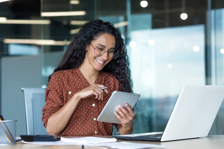 Smiling woman with curly hair and glasses working in a modern office, using a tablet and stylus while seated at a desk with a laptop and papers.