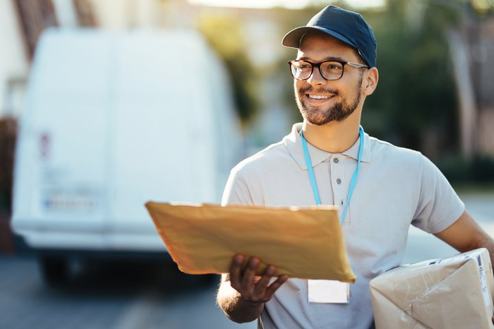 Smiling delivery man wearing a cap and glasses, holding a padded envelope and a cardboard box, standing in front of a white van on a sunny day.