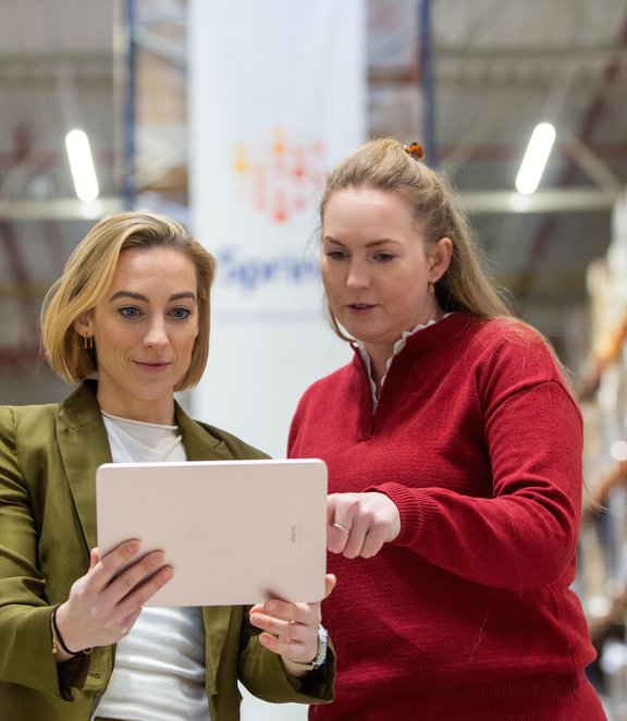 Two logistics professionals reviewing information on a tablet inside a warehouse aisle.