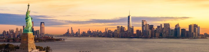 The Statue of Liberty in the foreground with the New York City skyline and One World Trade Center illuminated by a warm sunset in the background.