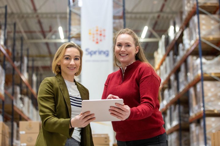 Two smiling women in a warehouse, holding a tablet.