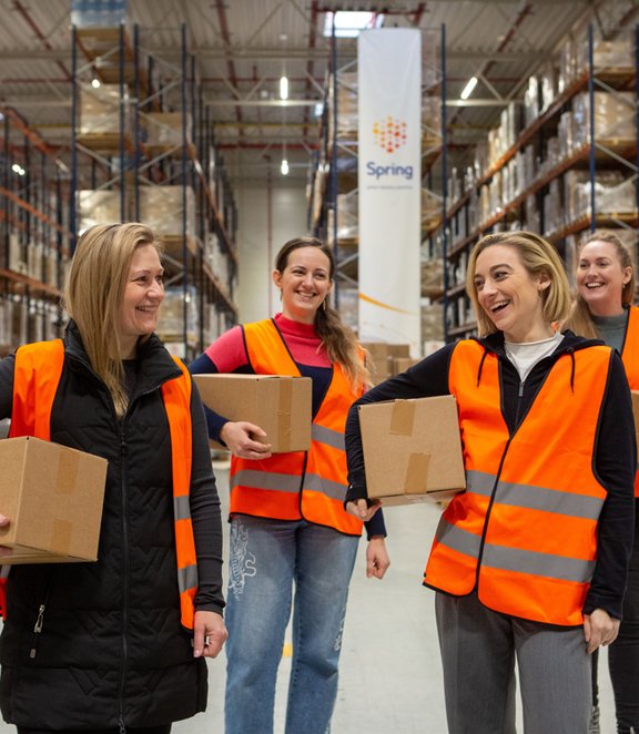 4 smiling women in reflective vests hold parcels inside a warehouse.