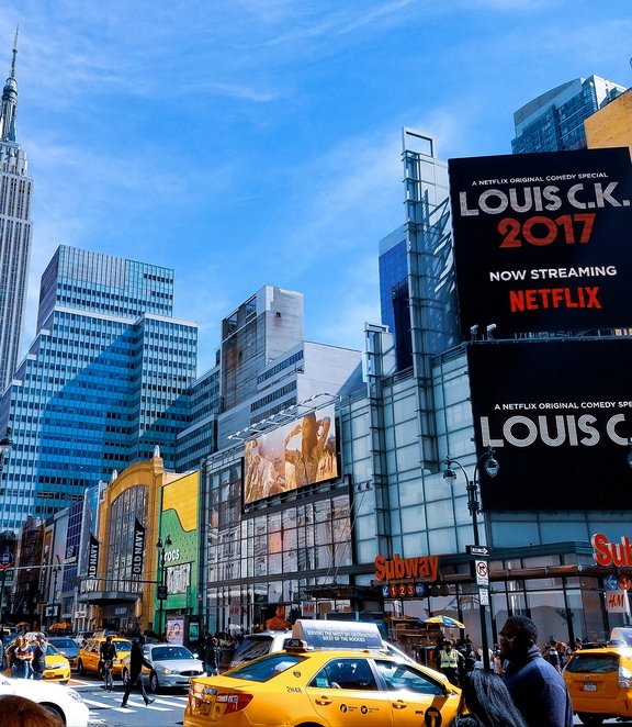 usy New York City street scene featuring yellow taxis, a Subway entrance, and a large Netflix billboard for Louis C.K.'s 2017 comedy special.